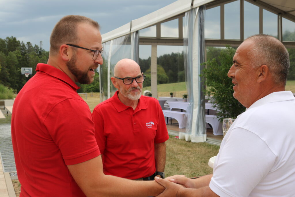 Three men in red shirts conversing in a grassy area with dining tables in the background