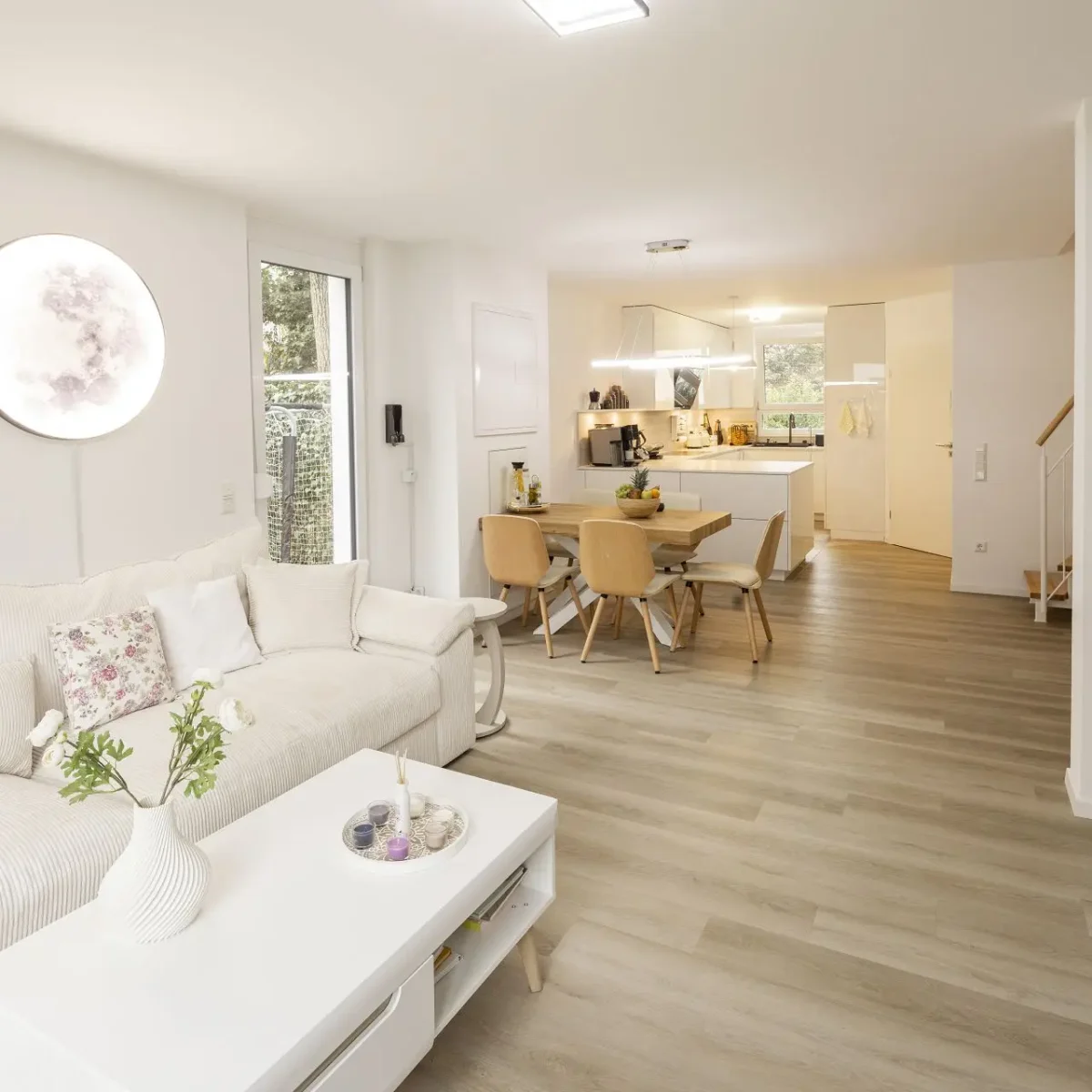 A clean and spacious living room with a white color scheme, featuring a couch, dining table, and kitchen area